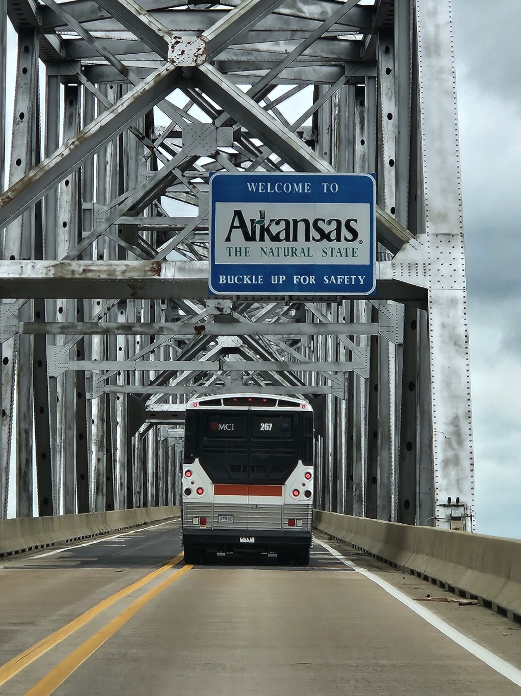 Une route qui passe sur un pont mais presque toute l'image est prise par la ferraille sur le pont. Sur le coté droit un peu de grisaille. Un bus est devant nous et sur la feraille du pont : Un panneau Arkansas